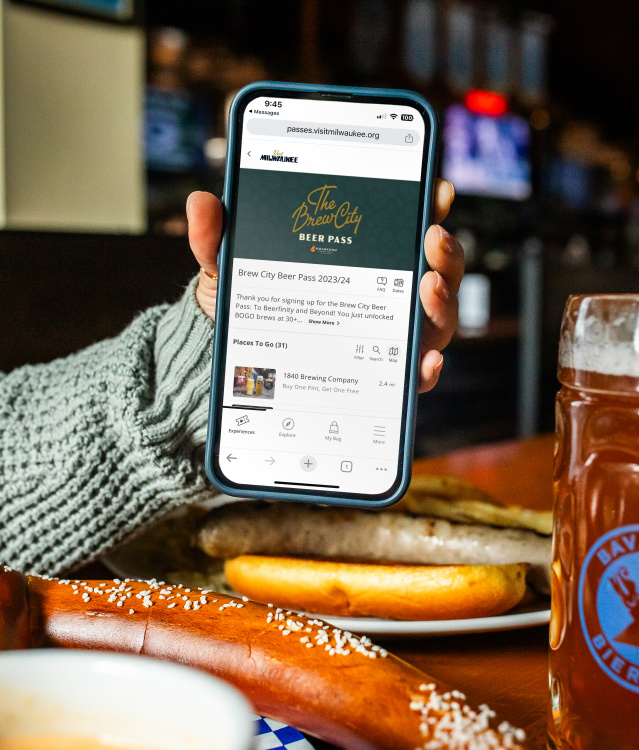 a hand holding up a phone displaying the Brew City Beer Pass over a table with a Bavarian Pretzel and a large pint of beer
