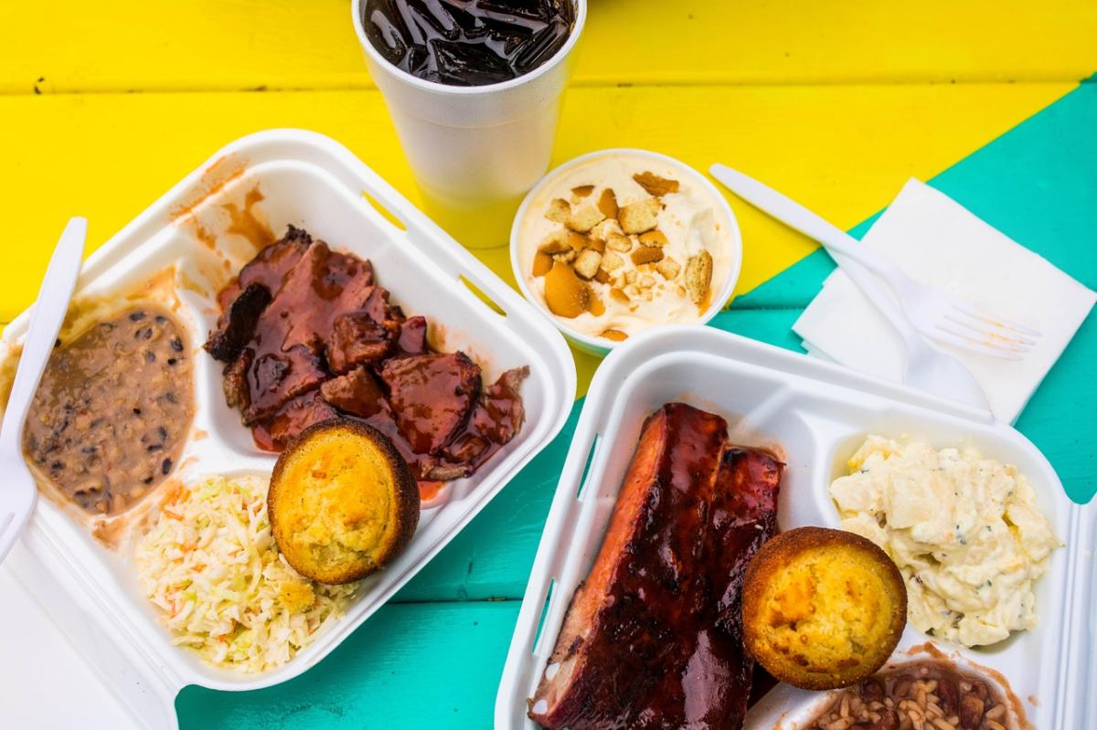 Two take away containers loaded with BBQ ribs and sides on a brightly colored table.