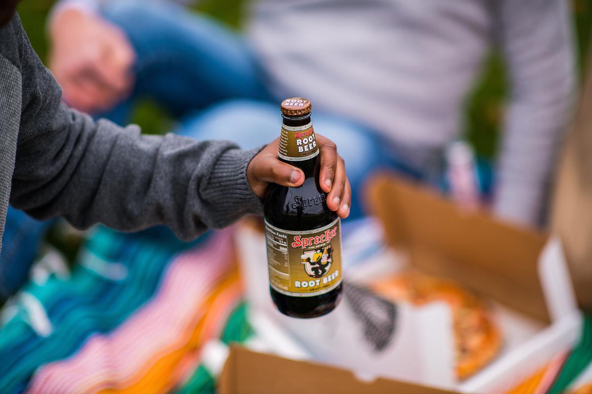 Person holding a Sprecher Root Beer bottle over a multicolored blanked and pizza during a picnic