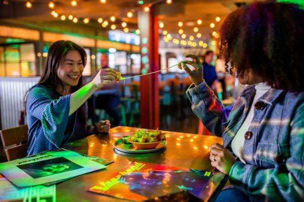 two women sitting at a table in a dim, string-light-lit bar pulling apart a cheese curd to stretch the cheese inside