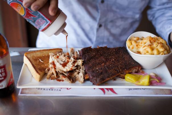 Person pouring BBQ sauce on a plate of of BBQ chicken and ribs with a side of mac and cheese.