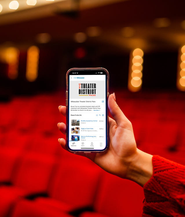 Hand holding up cellphone displaying homescreen of Milwaukee Theater District Pass in front of blurry theater background