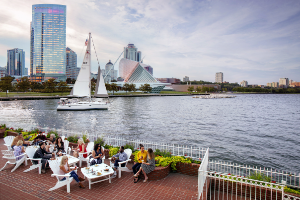a group of people eating on the patio in front of the Milwaukee skyline with a sailboat in the harbor